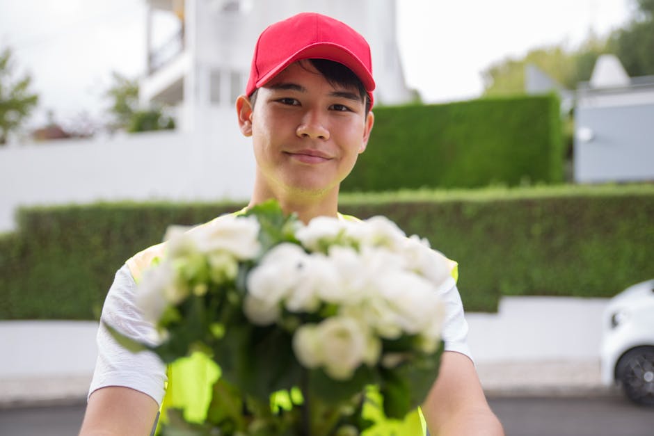 A young delivery person wearing a red cap and a high-visibility yellow vest holds a bouquet of fresh white flowers, including roses and other blooms, with green foliage. The flowers are arranged in a 