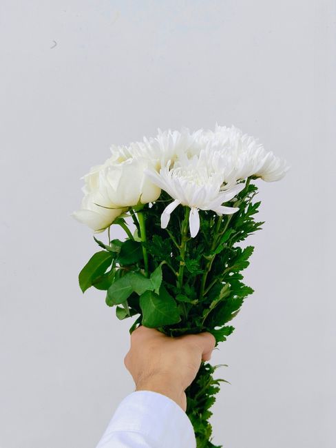 A hand holding a bouquet of fresh white flowers, including white roses and large white chrysanthemums, arranged with lush green foliage. The bouquet is wrapped in simple green stems, and the flowers a
