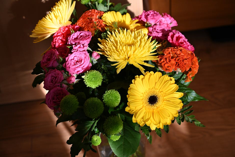 A vibrant floral bouquet arranged with large yellow gerbera daisies, pink and orange carnations, bright yellow chrysanthemums, and small green button chrysanthemums, all complemented by lush green fol