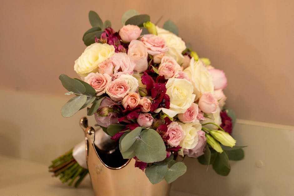A floral arrangement featuring a bouquet of fresh roses in soft pink and creamy white, complemented by deep burgundy flowers and lush green eucalyptus leaves. The bouquet is arranged in a rounded, nat