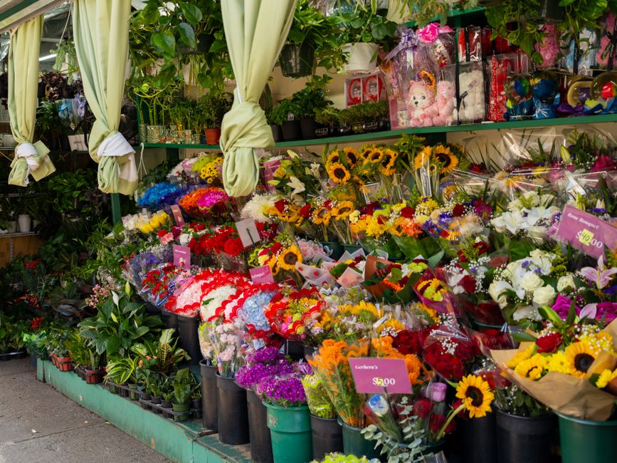 A vibrant floral display outside a florist's shop, showcasing a variety of fresh flower bouquets arranged on tiered shelves. The arrangement includes sunflowers with bright yellow petals and dark cent