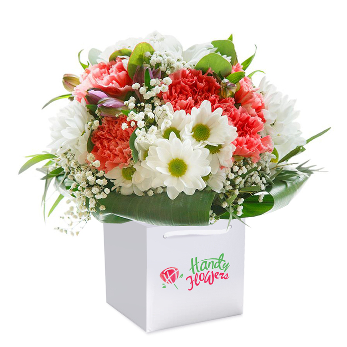 A masterfully arranged bouquet stands upright in a clean, square white gift bag, radiating freshness and vitality at the heart of Orpington. The rich blend of soft coral and peach carnations, with their dense, ruffled petals, establishes a warm and inviting mood, perfectly suited to the gentle pace of local life. Sprays of pure white daisies, each centered with lively yellow-green, are artfully interspersed-balancing the arrangement with a touch of cheerful brightness reminiscent of a morning stroll past Orpington's flower-laden market stalls. Wispy clouds of baby's breath delicately meander throughout, lightening the visual weight and creating a sense of movement. Notably, a cluster of closed buds tinged with purple hues peeks through on one side, hinting at the promise of further bloom and depth within the composition. The entire bouquet is cradled by swathes of robust, deep green leaves and glossy foliage, forming an elegant natural wrap at the base, grounding the display with the deep greens characteristic of Orpington's woodland edges. The bag's white ribbon handles not only complement the design but speak to the practicality and intent behind local flower delivery-perfect for gifting along Sevenoaks Road or as a thoughtful surprise for a loved one across Orpington. Every stem and blossom has been placed with intention, reflecting the dedication of florists who believe every celebration, birthday, or heartfelt gesture deserves beauty as memorable as the community itself.