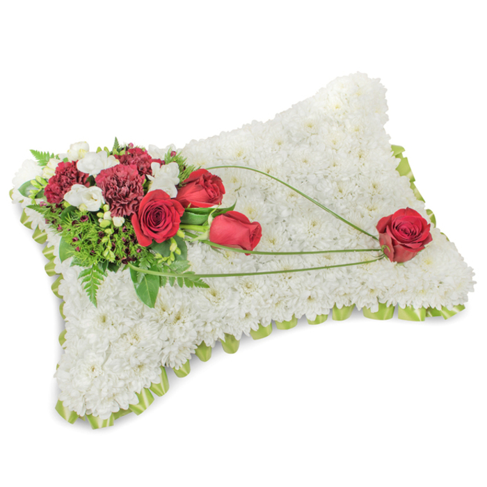 A beautifully balanced rectangular pillow of flowers, designed to convey solace and respect, sits composed against a neutral backdrop. The whole pillow form is lovingly built from hundreds of small, pure white chrysanthemums, packed so closely that their tiny petals form a sumptuous, fleece-like surface that invites calm and reflection. Encircling the gently rounded edges, a pale lime-green ribbon or slender foliage peeks through the white florals, creating a delicate frame that softens the silhouette. Anchoring the arrangement on the upper-left corner is a lush spray of deep crimson: velvety red roses at varying stages of bloom, rich burgundy carnations, and clusters of petite white freesias or spray chrysanthemums that introduce lightness and subtle scent. Behind and between the blooms, layers of greenery-soft fern fronds and textured leaves-add depth and a natural, verdant backdrop. A trio of elegant, bright-green bear-grass strands arcs from the main cluster and traces a graceful diagonal across the chrysanthemum field, culminating in a solitary red rose positioned on the lower right; this single bloom feels intentionally placed, a quiet emblem of love and remembrance. The interplay of textures-smooth satin ribbon, plush chrysanthemums, silky rose petals, and ruffled carnations-creates visual richness while remaining subdued and respectful. In Orpington settings such as Goddington Park memorials or chapel tributes along Sevenoaks Road, this kind of cushion is chosen for its dignified simplicity and ability to communicate heartfelt sympathy without words.