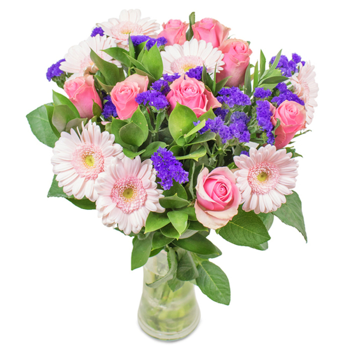 An elegantly arranged floral bouquet sits in a clear glass vase filled with fresh water, set against a crisp, white background-symbolic of Orpington's bright community spirit. At its heart, seven to nine pink roses display their classic spiral centers, some tightly furled while others gently opening, showing gradients from the softest petal pink to a richer rose blush. Interwoven among these are five to seven pale pink Gerbera daisies, recognizable by their wide, round faces and subtle cream-toned centers, reminiscent of the springtime blooms seen in local parks like Priory Gardens. Clusters of vivid purple statice are scattered throughout, their airy texture and deep colour providing a lively, modern twist. Lush green foliage, abundant and glossy, forms a generous base that both frames and refreshes the arrangement, evoking walks under the leafy canopies near Goddington Park. The composition is bright yet soothing, each element arranged with an eye for balance and natural movement. This bouquet is versatile-ideal for celebrating birthdays, anniversaries, or new arrivals in Orpington, and well-suited for brightening any living space or professional setting. It embodies the thoughtful artistry of local florists, every stem selected to convey both joy and affection with a personal, neighbourhood touch.
